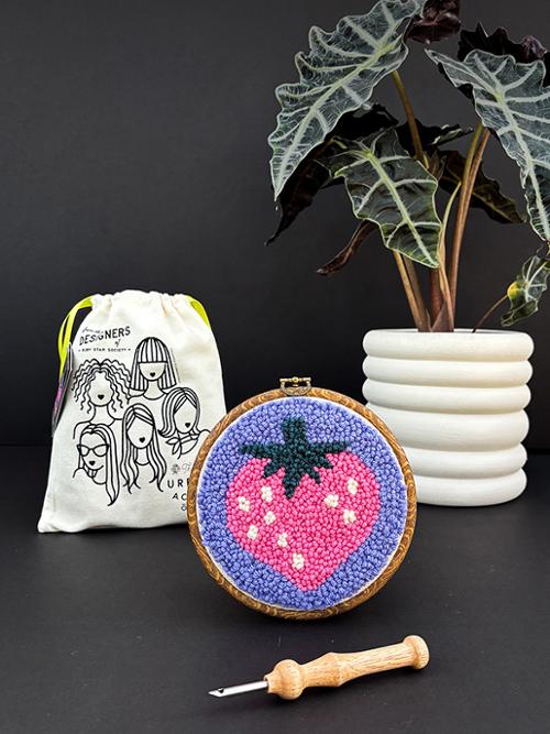 Embroidery hoop with strawberry design, white drawstring bag, and plant on dark background