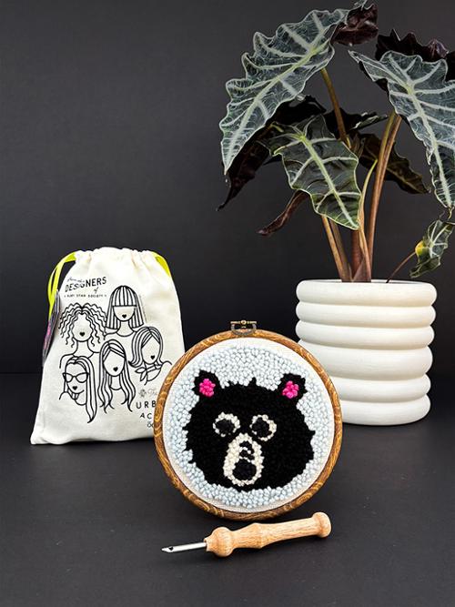 Embroidery hoop with a black and white bear design, surrounded by a drawstring bag and plant on a dark surface.